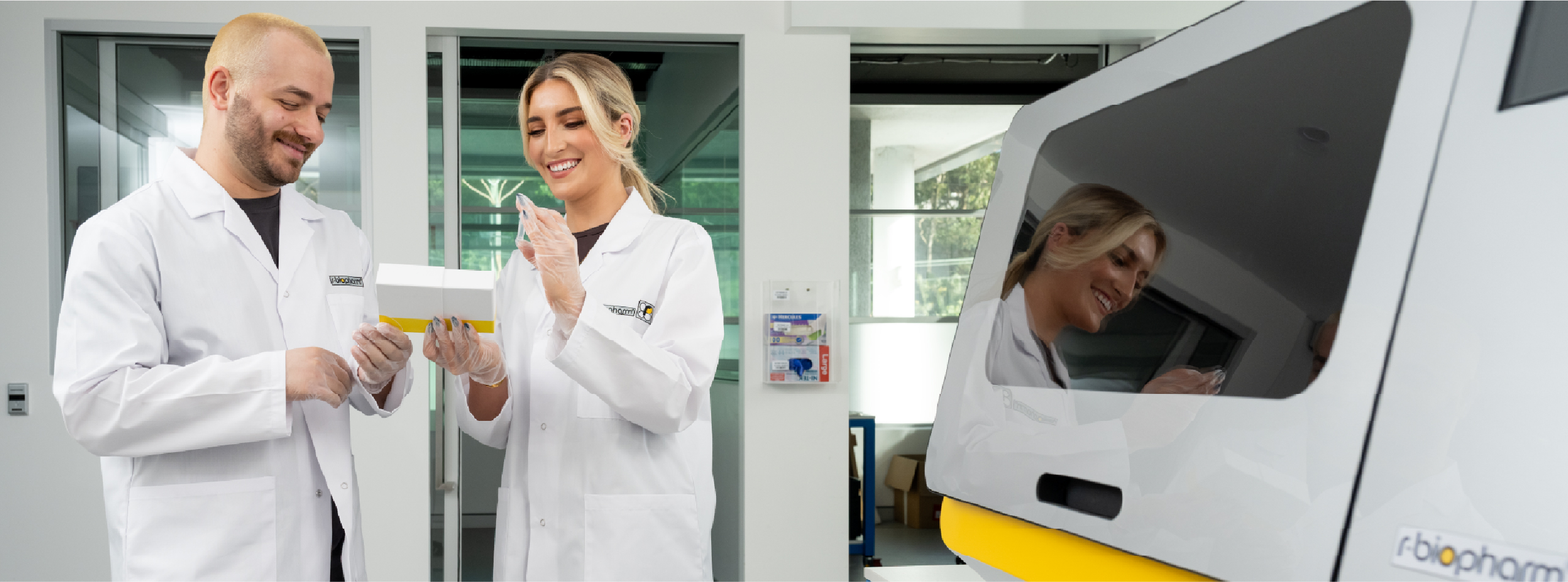 Two individuals in lab coats stand beside a white and yellow machine in a laboratory setting.