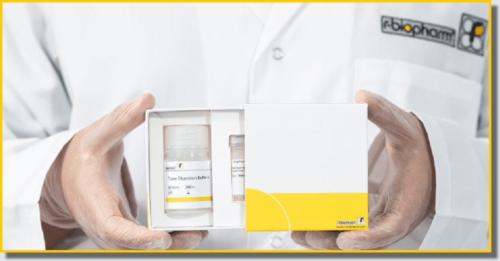 A man in a lab coat holds a box with a yellow and white label, standing in a laboratory setting.