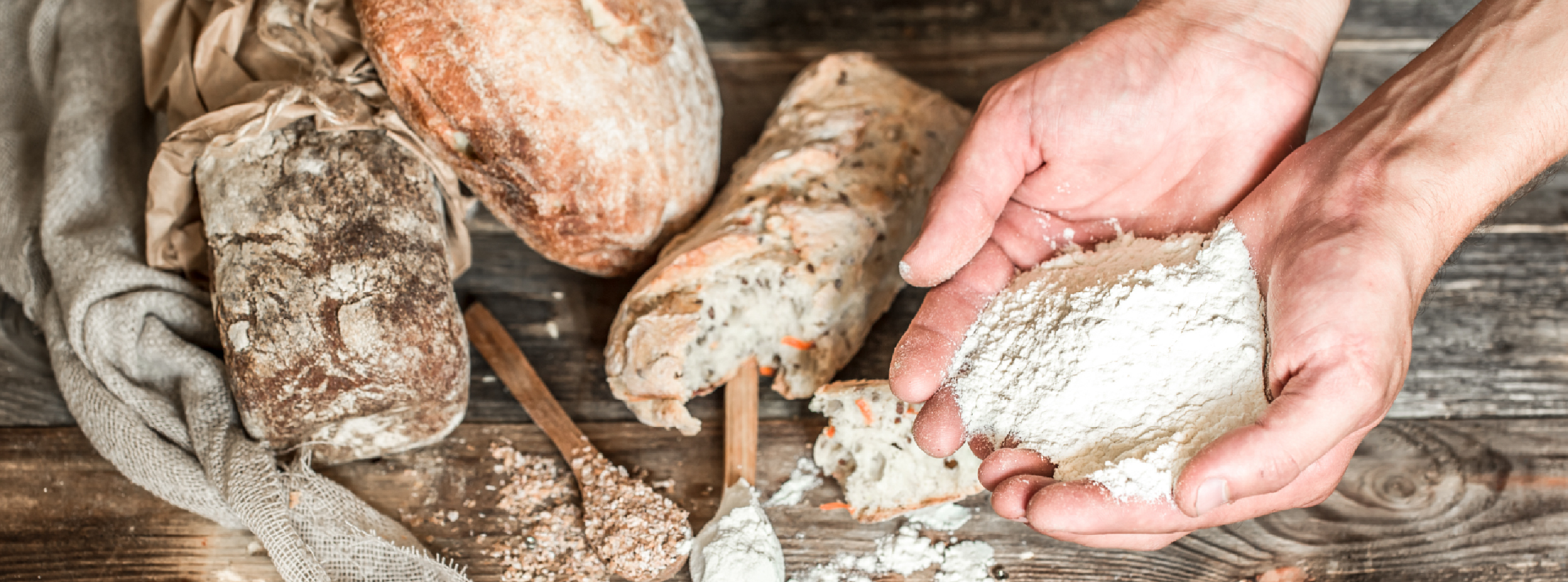 Hands holding flour and a loaf of bread on a rustic wooden table, emphasising the connection between ingredients and baking.