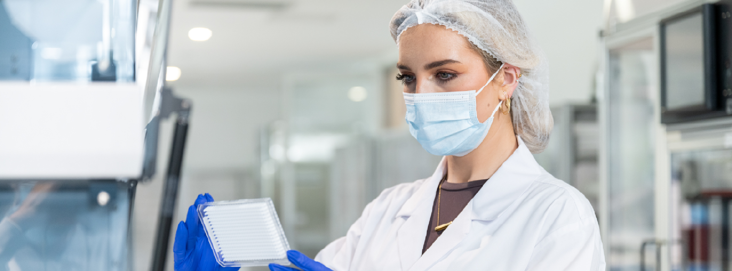A woman in a lab coat, mask and hairnet holds a test panel, focused on her scientific work in a laboratory setting.