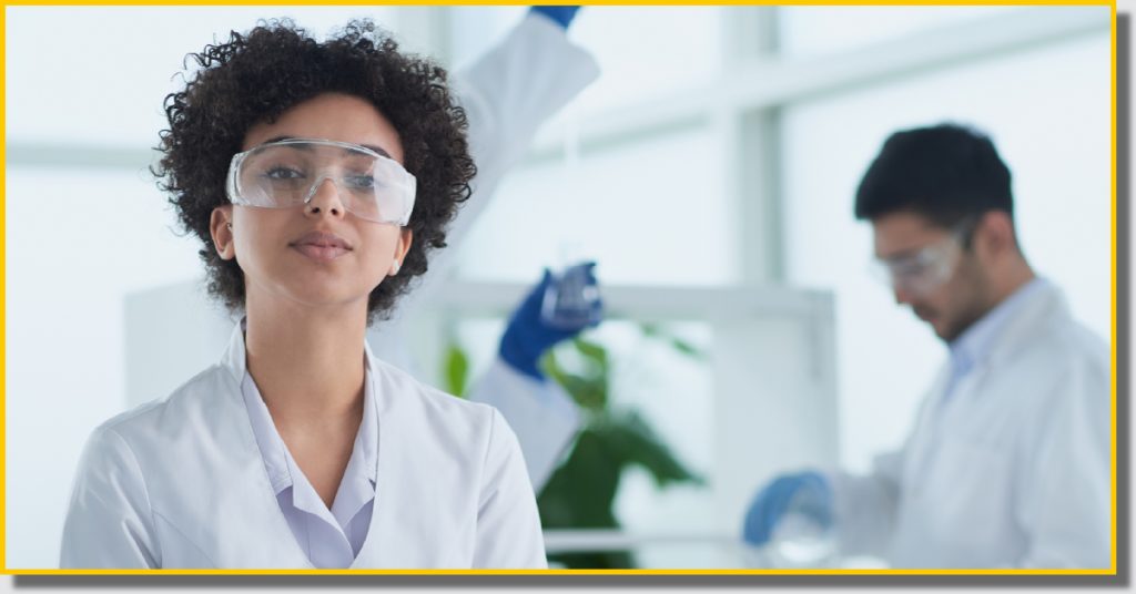 A woman in a lab coat and goggles stands next to a man in a lab coat