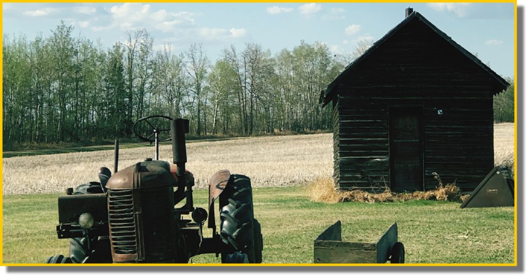 An old tractor beside a small cabin, set in the middle of a vast green field under a clear blue sky.