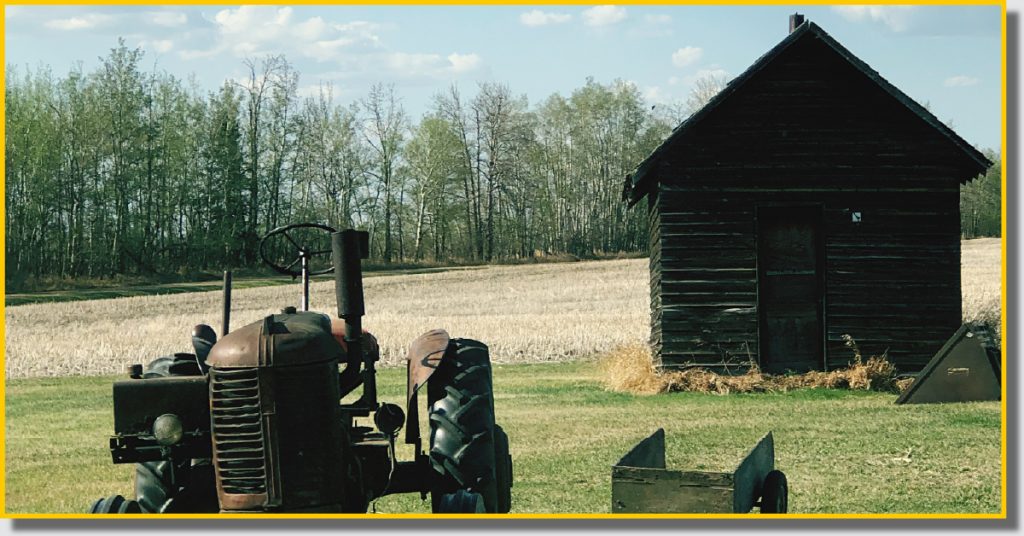 An old tractor beside a small cabin, set in the middle of a vast green field under a clear blue sky.