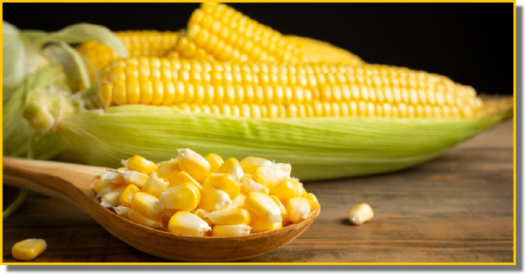 A close-up of yellow corn kernels beside a wooden spoon on a rustic surface.
