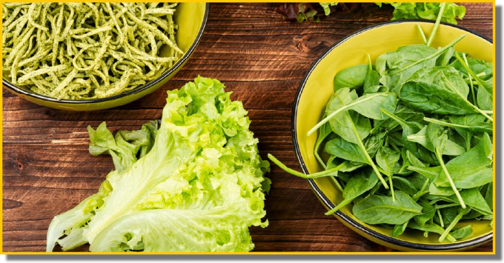 A bowl of green pasta with fresh spinach next to a bowl of crisp lettuce