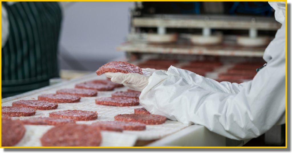 A worker in a white coat places hamburgers onto a conveyor belt in a food production facility.
