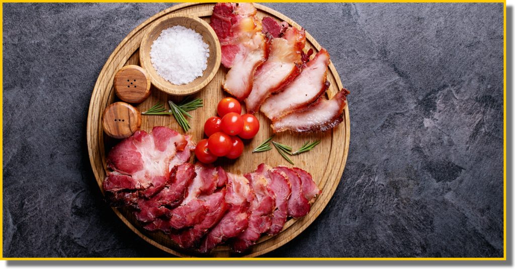 A wooden platter displaying various pork cuts, fresh, ripe tomatoes, salt, pepper and rosemary