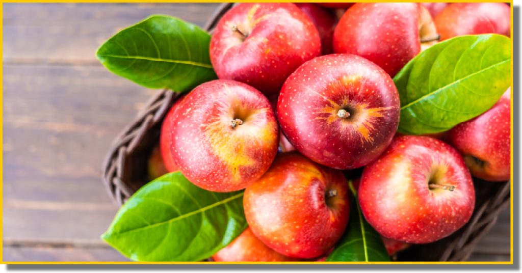 A basket filled with red apples topped with green leaves set on a brown table
