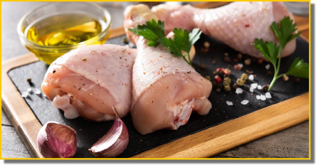 Raw chicken legs on a cutting board, accompanied by garlic cloves and a drizzle of oil.