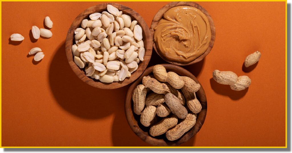 Two bowls of peanuts, one with shells and one without, next to a jar of creamy peanut butter on a wooden surface.