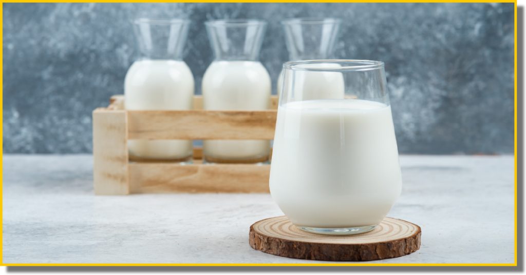 A glass of milk placed on a wooden coaster with 3 bottles of milk in the background