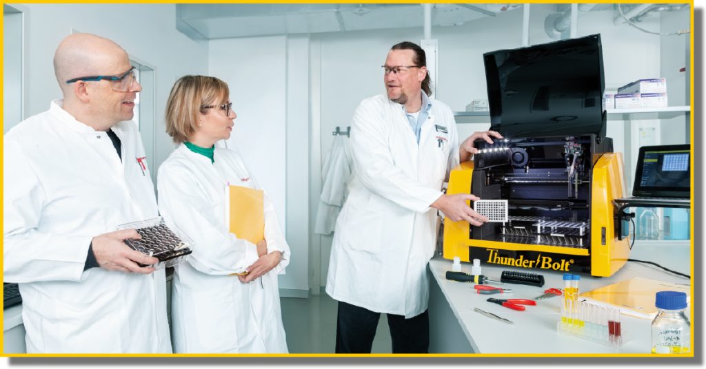 Three individuals in lab coats examining a complex machine in a laboratory setting.