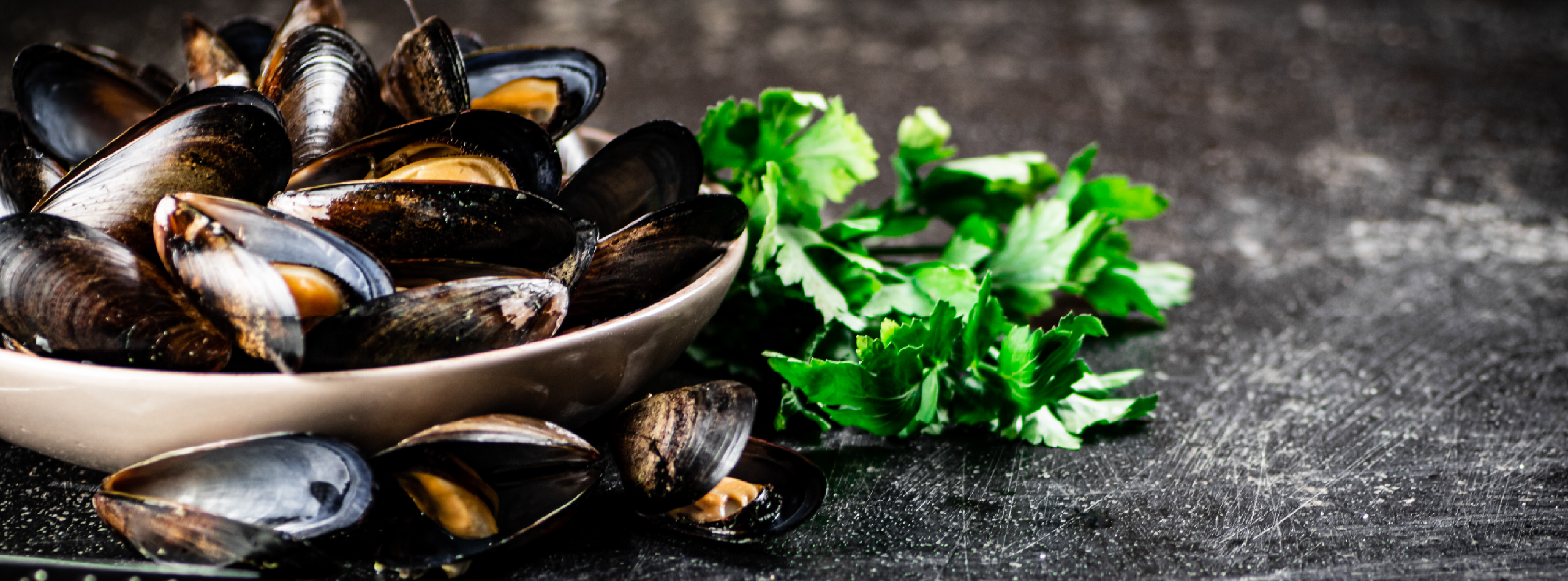 A bowl of mussels garnished with parsley sits on a dark table