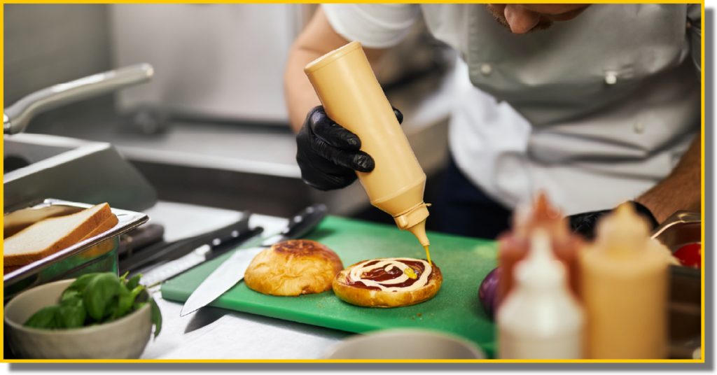 A chef prepares a burger on a green cutting board, surrounded by fresh ingredients and kitchen tools.