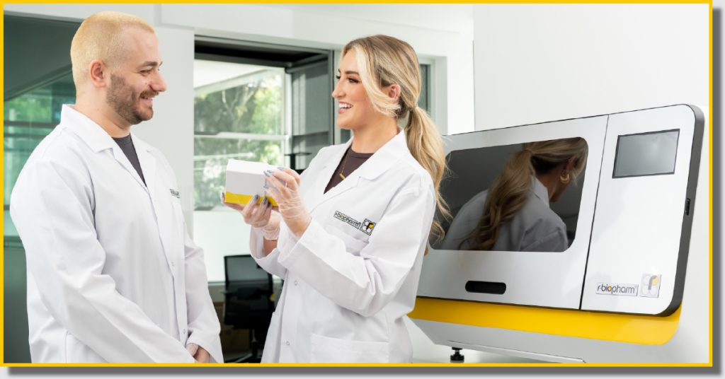 Two individuals in lab coats stand beside a scientific machine in a laboratory setting.