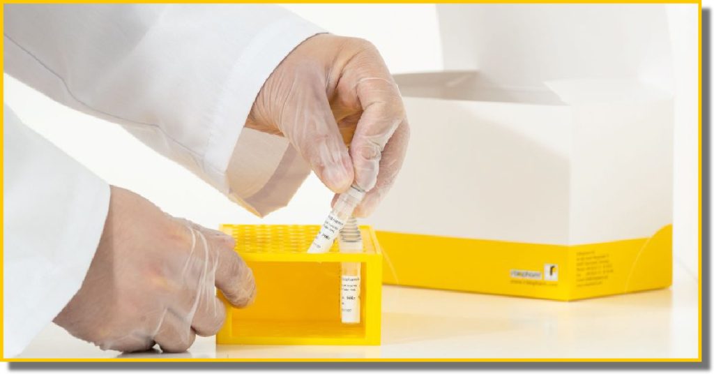 a close of a man in a lab coat's hands inside clear gloves handling tubes inside a yellow box next to product packaging